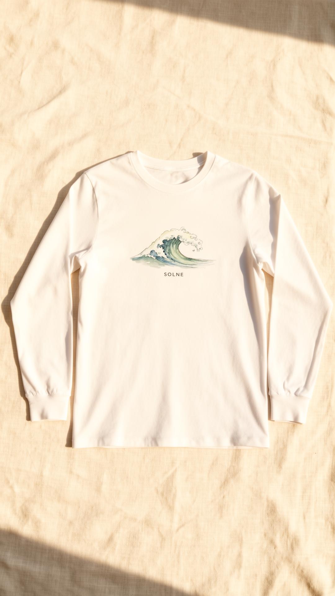 A white long-sleeve cotton tee with a hand-painted watercolor wave and the SOLNE wordmark, in warm afternoon light